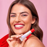 Woman smiling while holding a silver tube of Naked Sundays Candy Cane SPF50 lip treatment with a sheer red tint