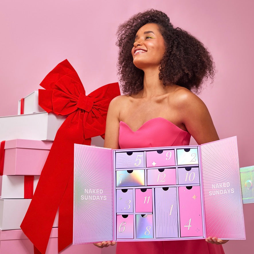 Woman holding a pink Naked Sundays 12 Days of Glow advent calendar with numbered drawers against a stack of gift boxes