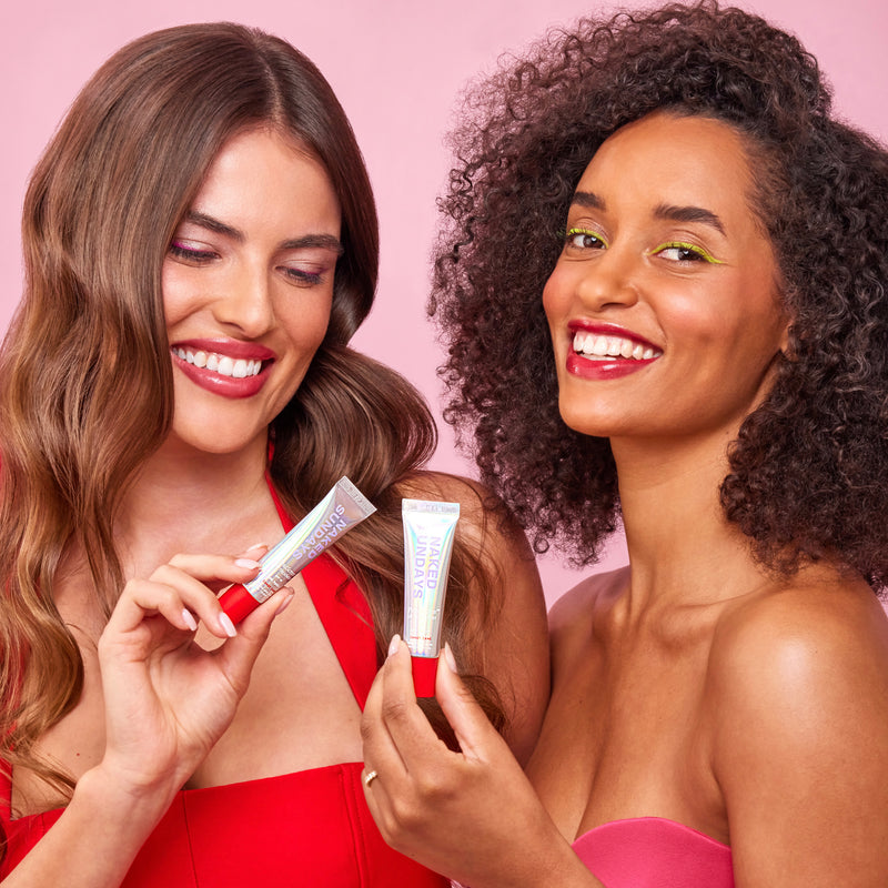 Two smiling women holding cosmetic tubes against a solid pink background.
