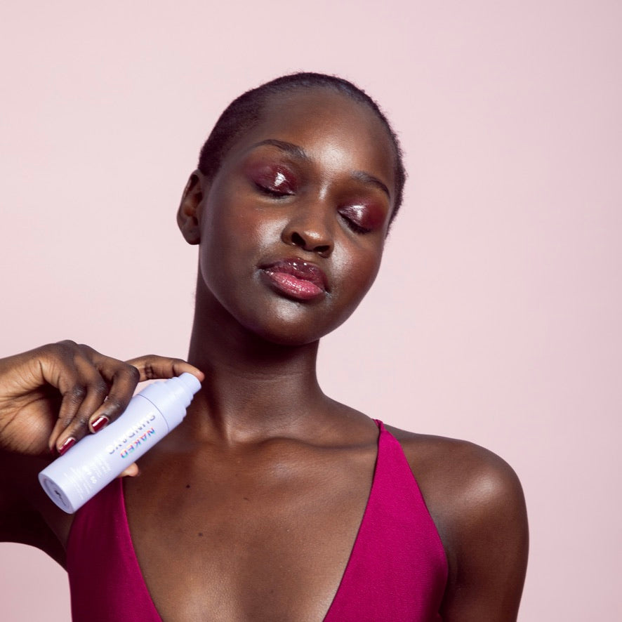 Woman applying a Naked Sundays Glow Mist SPF spray bottle to her neck against a pink background