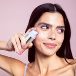Woman applying a swatch of Naked Sundays CabanaClear Illuminating Water Gel Serum SPF50 to her face against a pink background