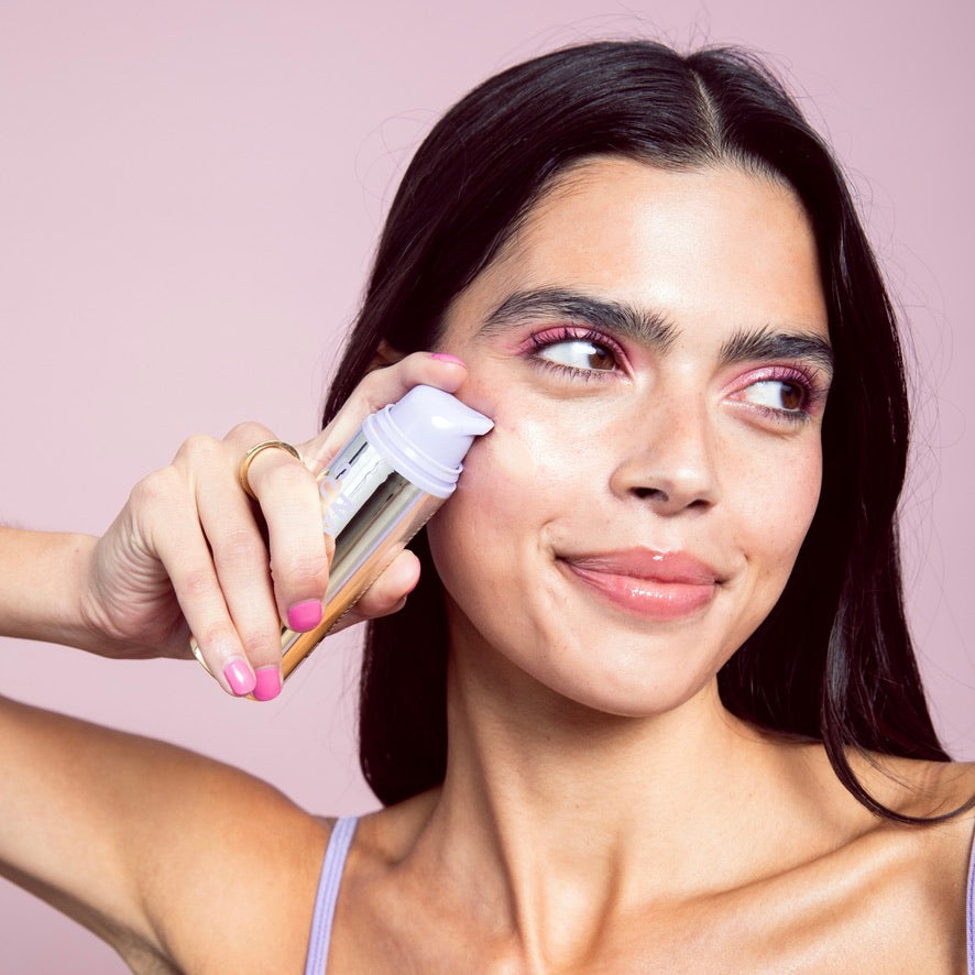 Woman applying a swatch of Naked Sundays CabanaClear Illuminating Water Gel Serum SPF50 to her face against a pink background