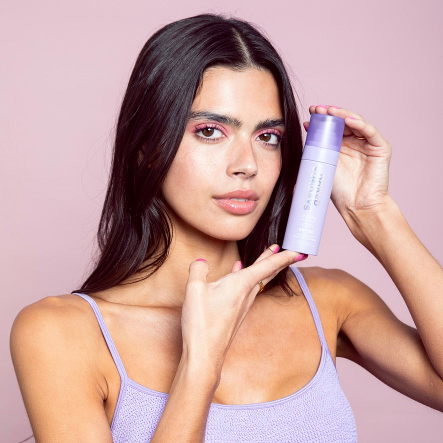 Woman holding Naked Sundays Glow Mist SPF50 against a pink background