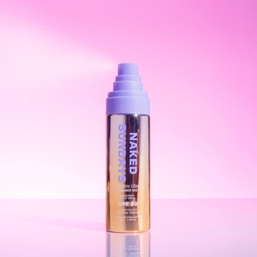 Bottle of Naked Sundays Golden Glow Shimmer Mist SPF50 sitting on a pink surface. The lid is off and the product is spraying in the left direction.