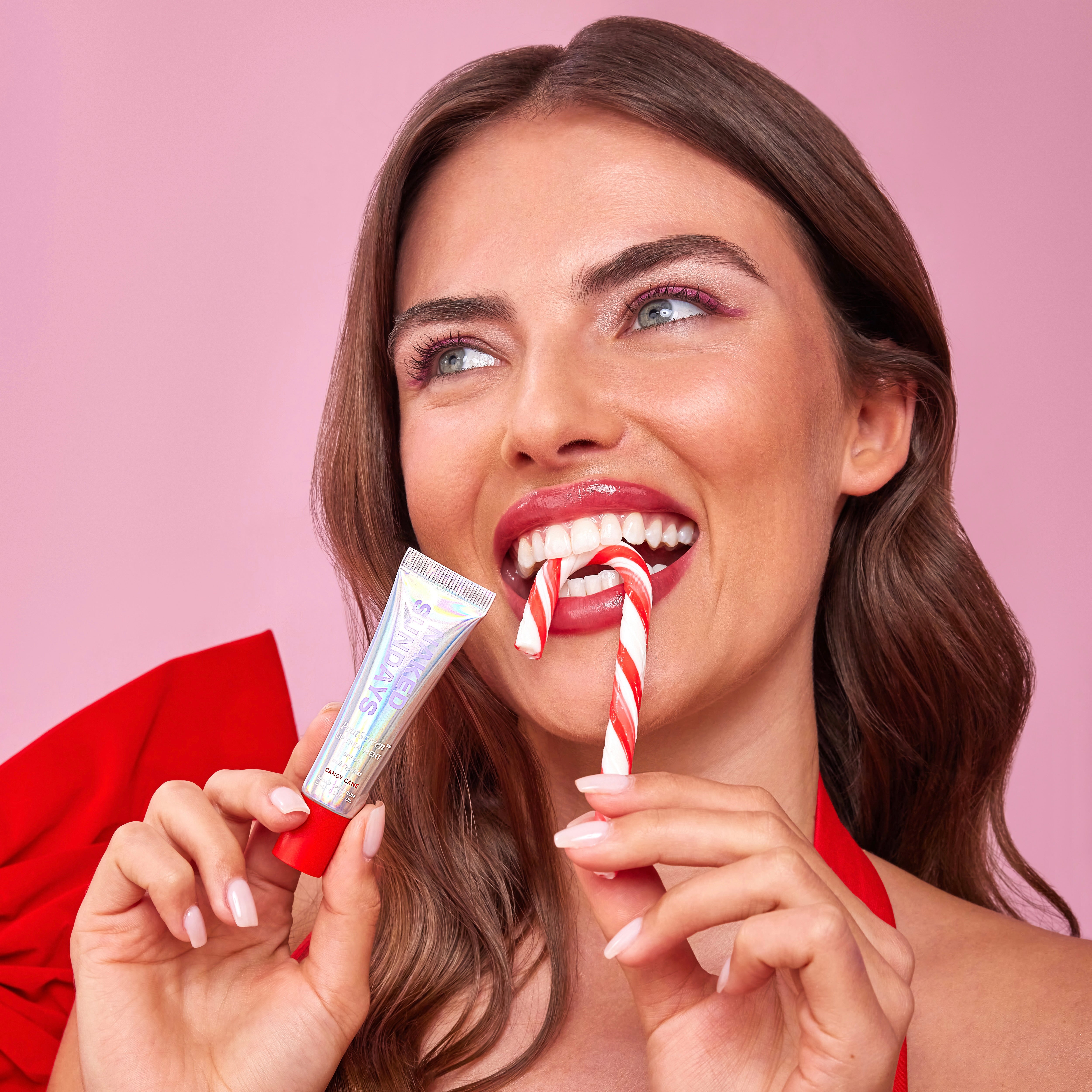 Woman holding a sheer red PoutScreen SPF50 lip treatment tube next to a candy cane against a pink background