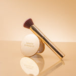 A gold makeup brush with dark brown bristles and a matching round compact mirror, both branded NAKED SUNDAYS, displayed on a reflective beige surface against a warm beige background
