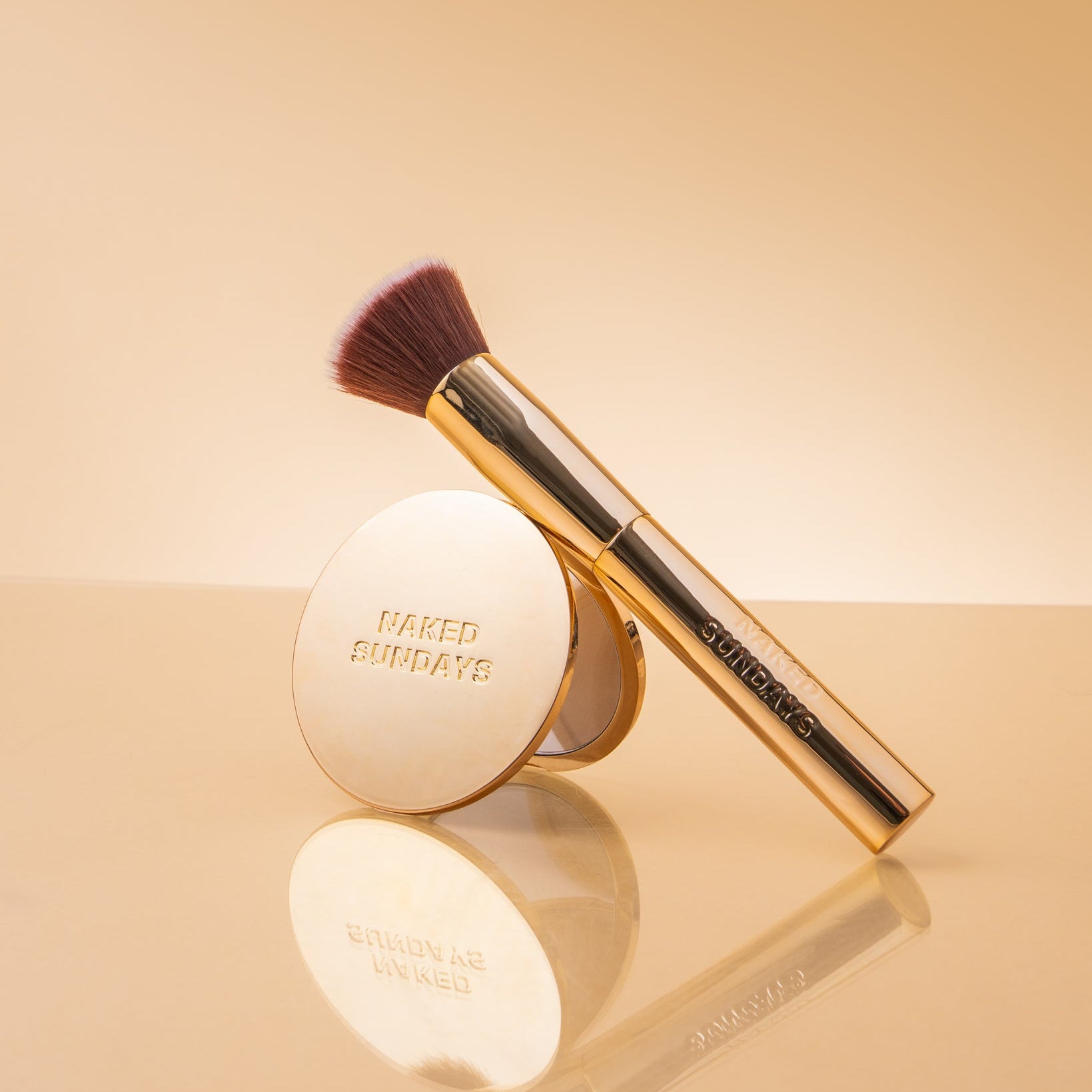A gold makeup brush with dark brown bristles and a matching round compact mirror, both branded NAKED SUNDAYS, displayed on a reflective beige surface against a warm beige background