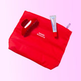 A red makeup bag, a red hair clip and a tube of Naked Sundays PoutScreen in shade Candy Cane on a purple gradient background