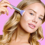 Woman applying makeup with a brush on a pink background