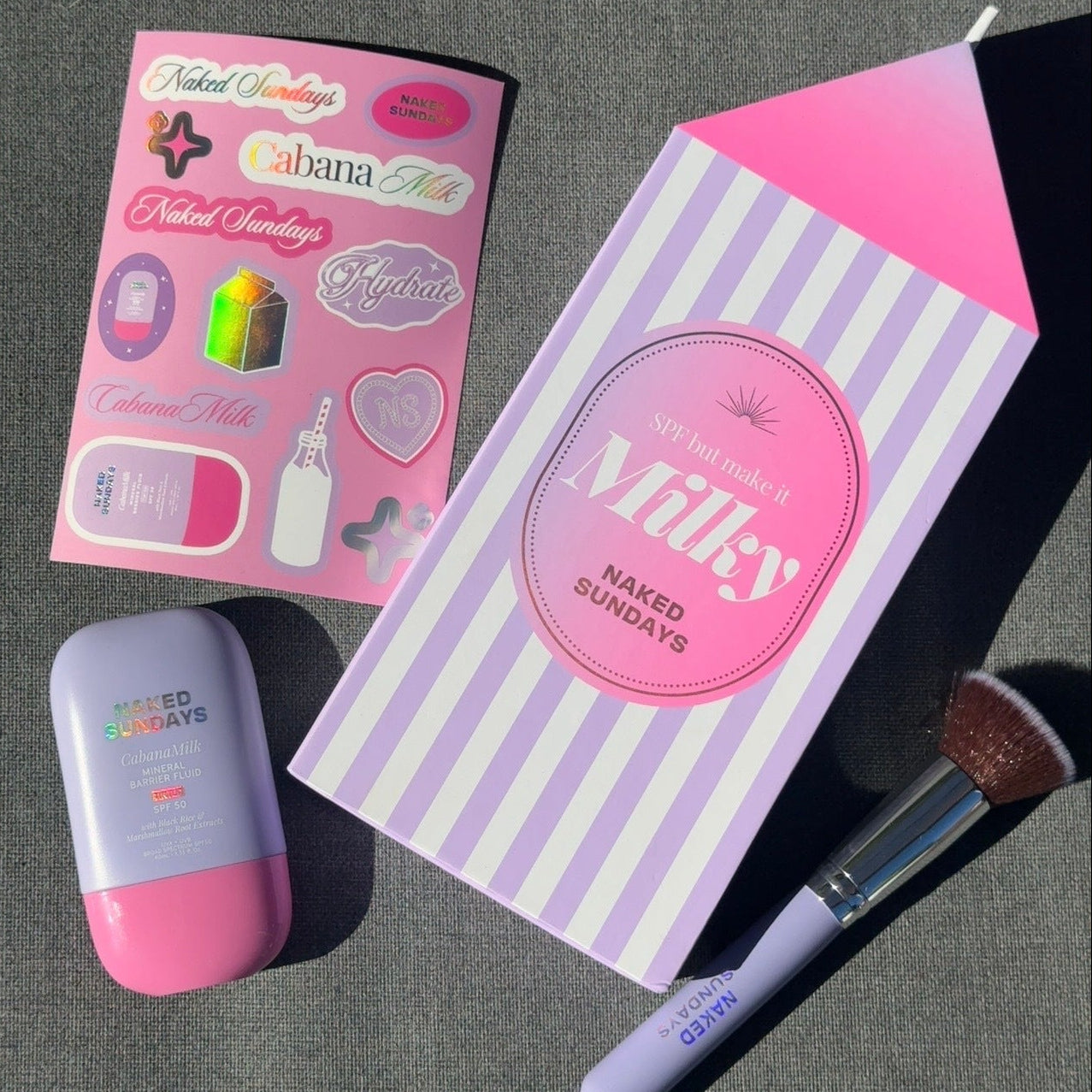 A styled flat lay of a beauty set on a dark gray textured surface, featuring a milk carton shaped box labeled 'Milky NAKED SUNDAYS', a pink and lavender makeup sponge, a pink and lavender compact, and a pink sticker sheet