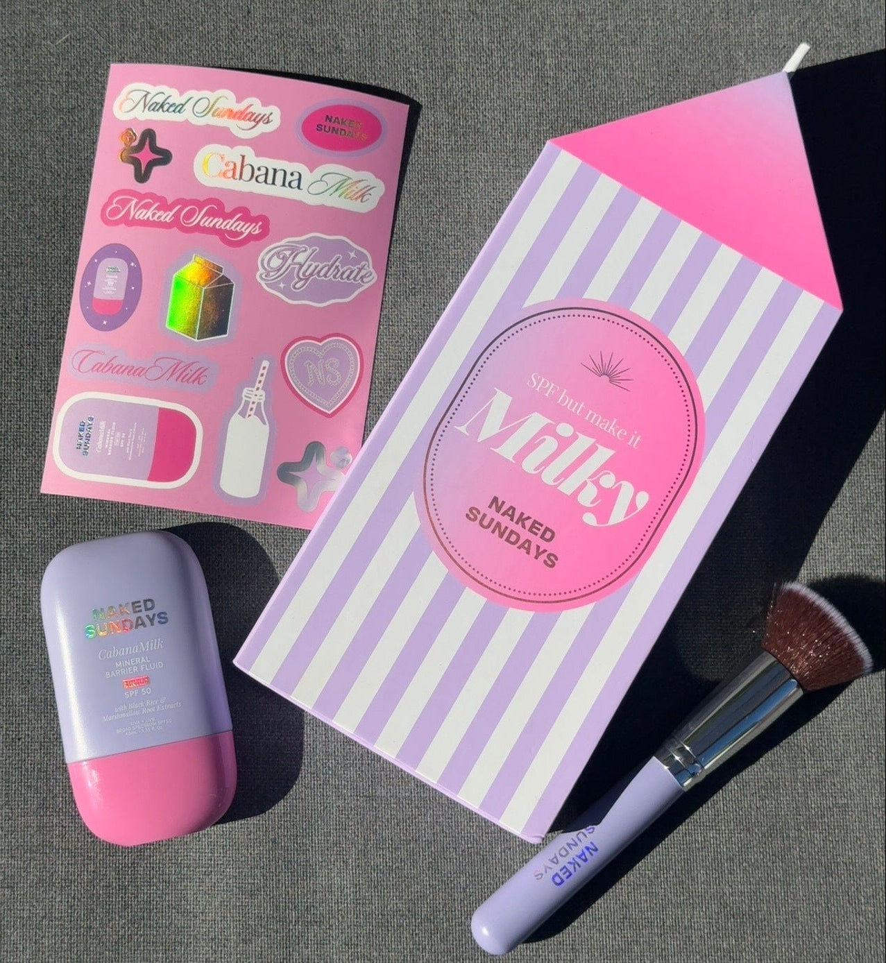 A styled flat lay of a beauty set on a dark gray textured surface, featuring a milk carton shaped box labeled 'Milky NAKED SUNDAYS', a pink and lavender makeup sponge, a pink and lavender compact, and a pink sticker sheet