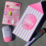 A styled flat lay of a beauty set on a dark gray textured surface, featuring a milk carton shaped box labeled 'Milky NAKED SUNDAYS', a pink and lavender makeup sponge, a pink and lavender compact, and a pink sticker sheet