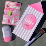 A styled flat lay of a beauty set on a dark gray textured surface, featuring a milk carton shaped box labeled 'Milky NAKED SUNDAYS', a pink and lavender makeup sponge, a pink and lavender compact, and a pink sticker sheet