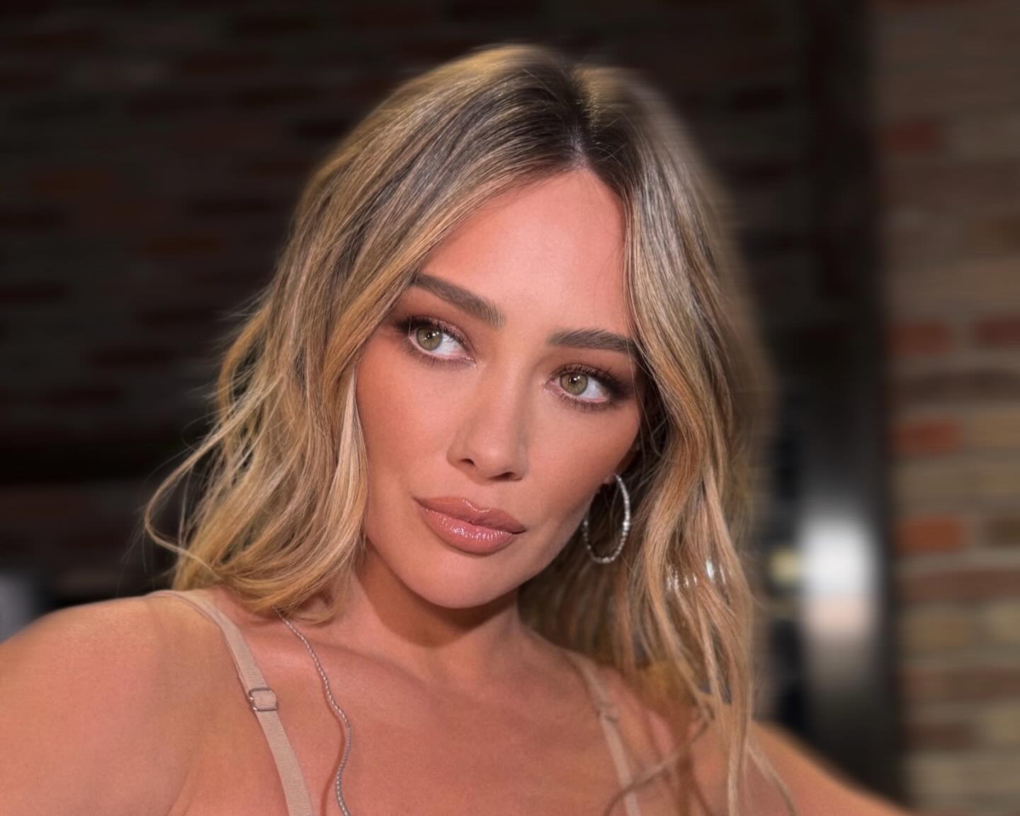 Woman with blonde hair and hoop earrings against a brick wall background