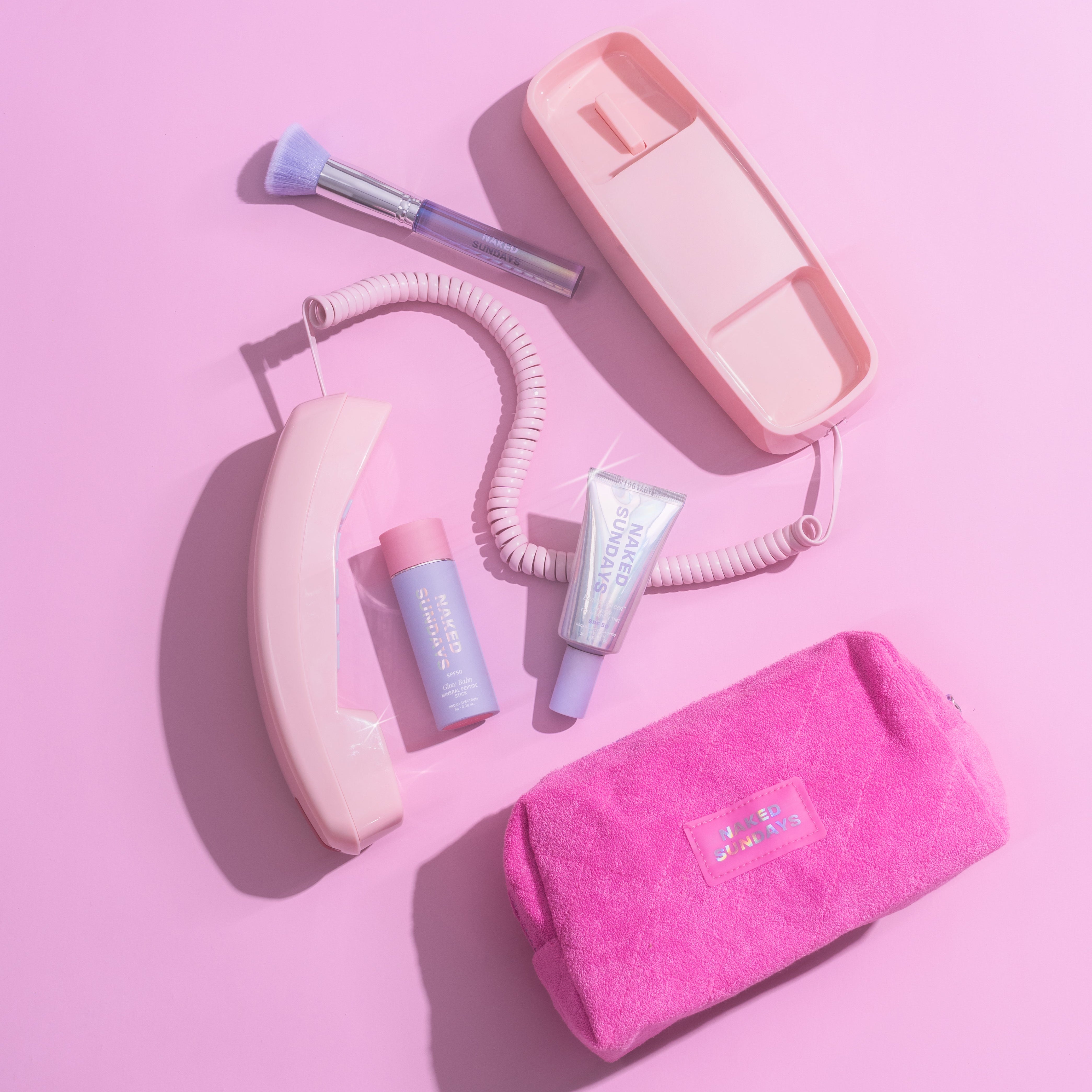 Pink makeup items including a brush, phone, and pouch on a pink background