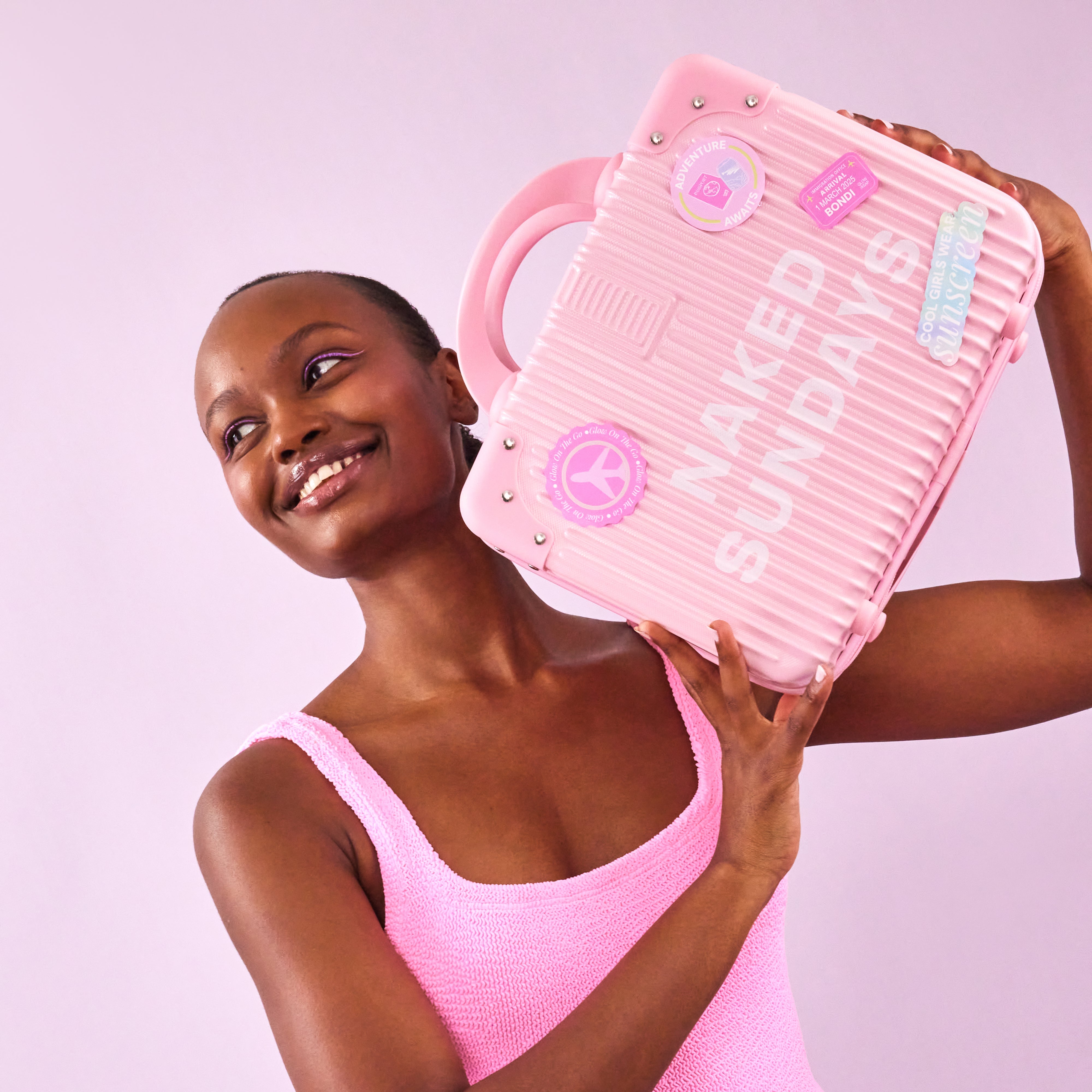 Woman in a pink swimsuit holding a pink bag with Naked Sundays branding against a light purple background