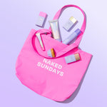 Pink tote bag with 'Naked Sundays' text containing beauty products on a light purple background