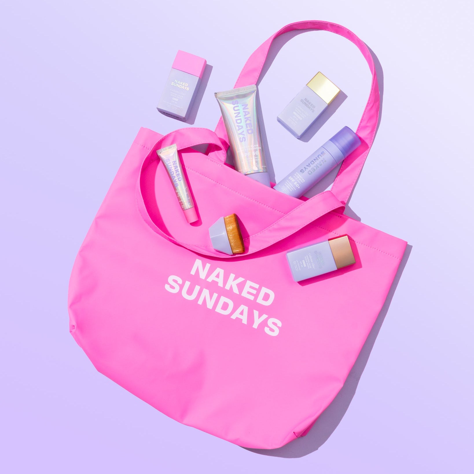 Pink tote bag with 'Naked Sundays' text containing beauty products on a light purple background
