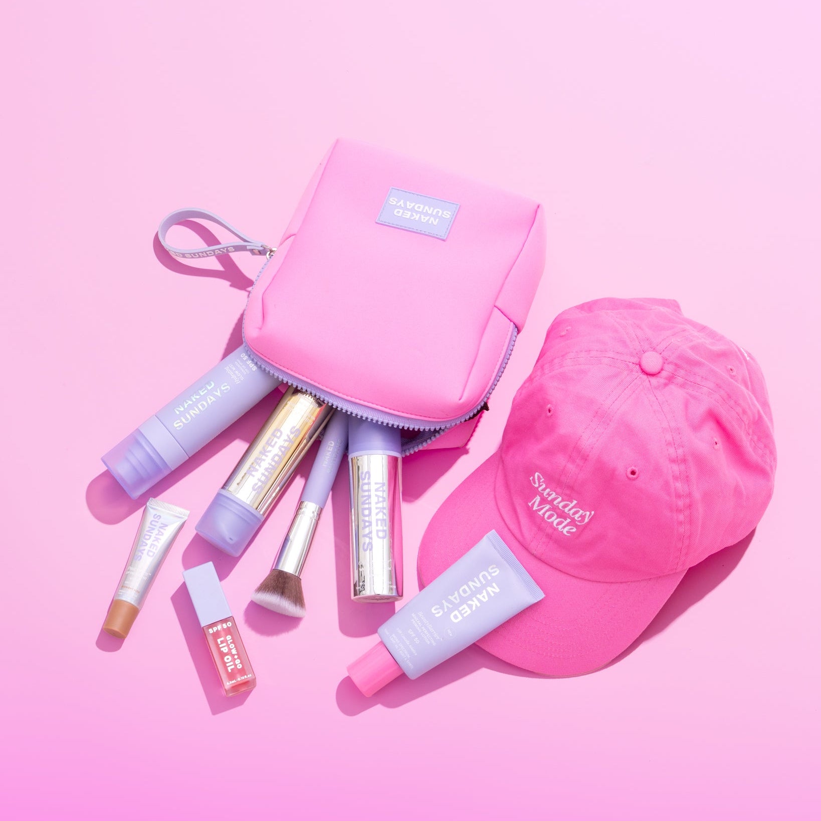 A pink zippered cosmetic pouch with a small blue label, surrounded by makeup items and a pink baseball cap on a pink background