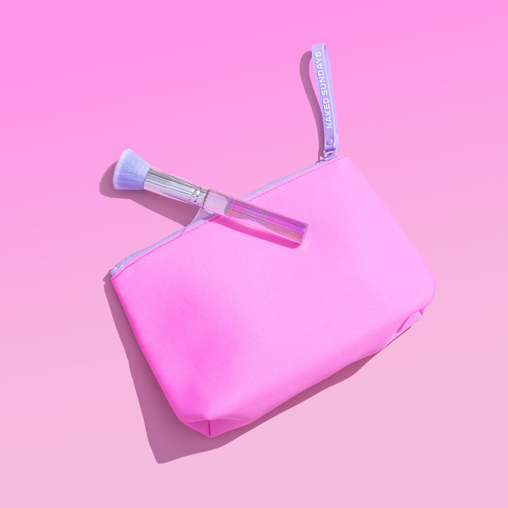 A bright pink cosmetic pouch with a matching pink zipper and handle, shown on a pink background with a makeup brush resting across the top