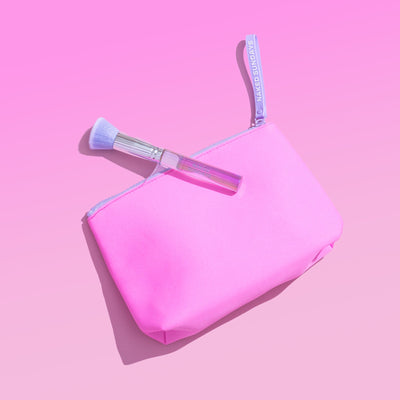 A bright pink cosmetic pouch with a matching pink zipper and handle, shown on a pink background with a makeup brush resting across the top.