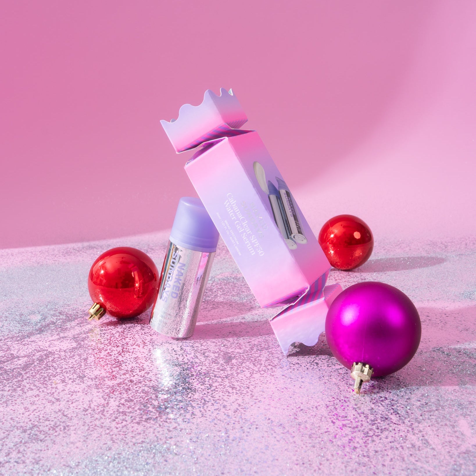 CabanaClear SPF50 water gel serum in a pink and blue holiday cracker, surrounded by red and purple ornaments on silver glitter