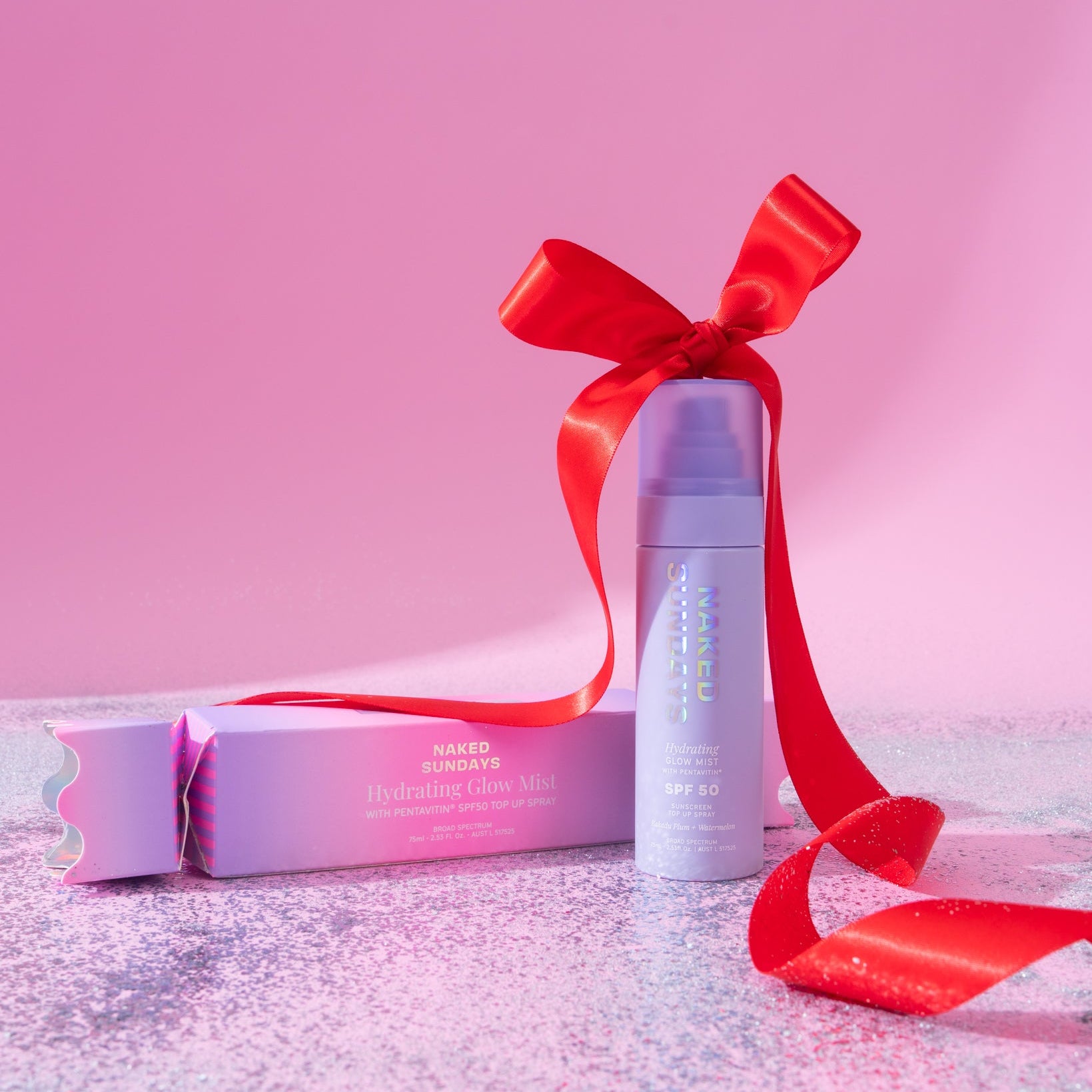 Naked Sundays SPF50 Hydrating Glow Mist in festive purple cracker packaging with a large red ribbon on a glittery surface