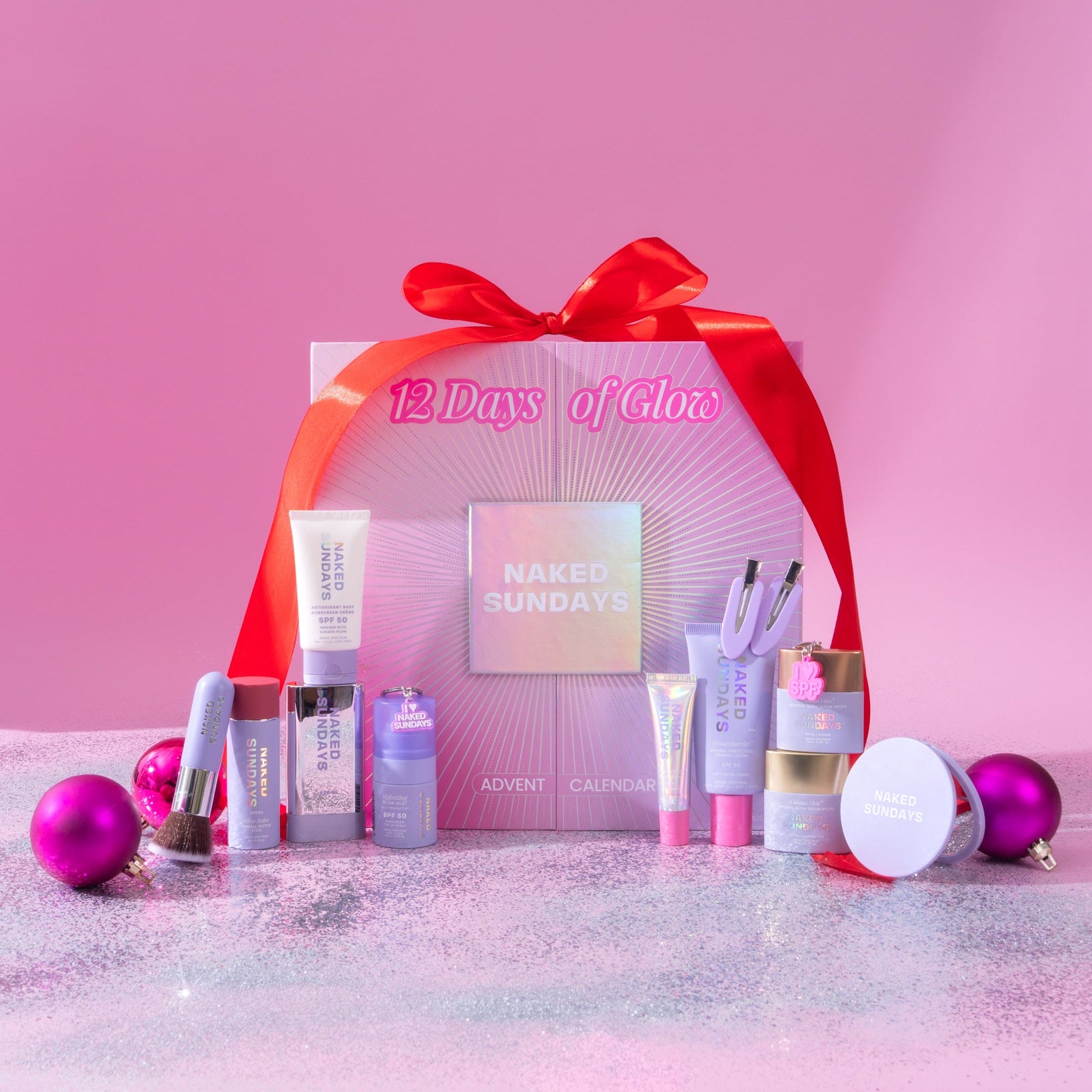 Naked Sundays 12 Days of Glow advent calendar box with red ribbon, surrounded by SPF skincare products and accessories on glitter