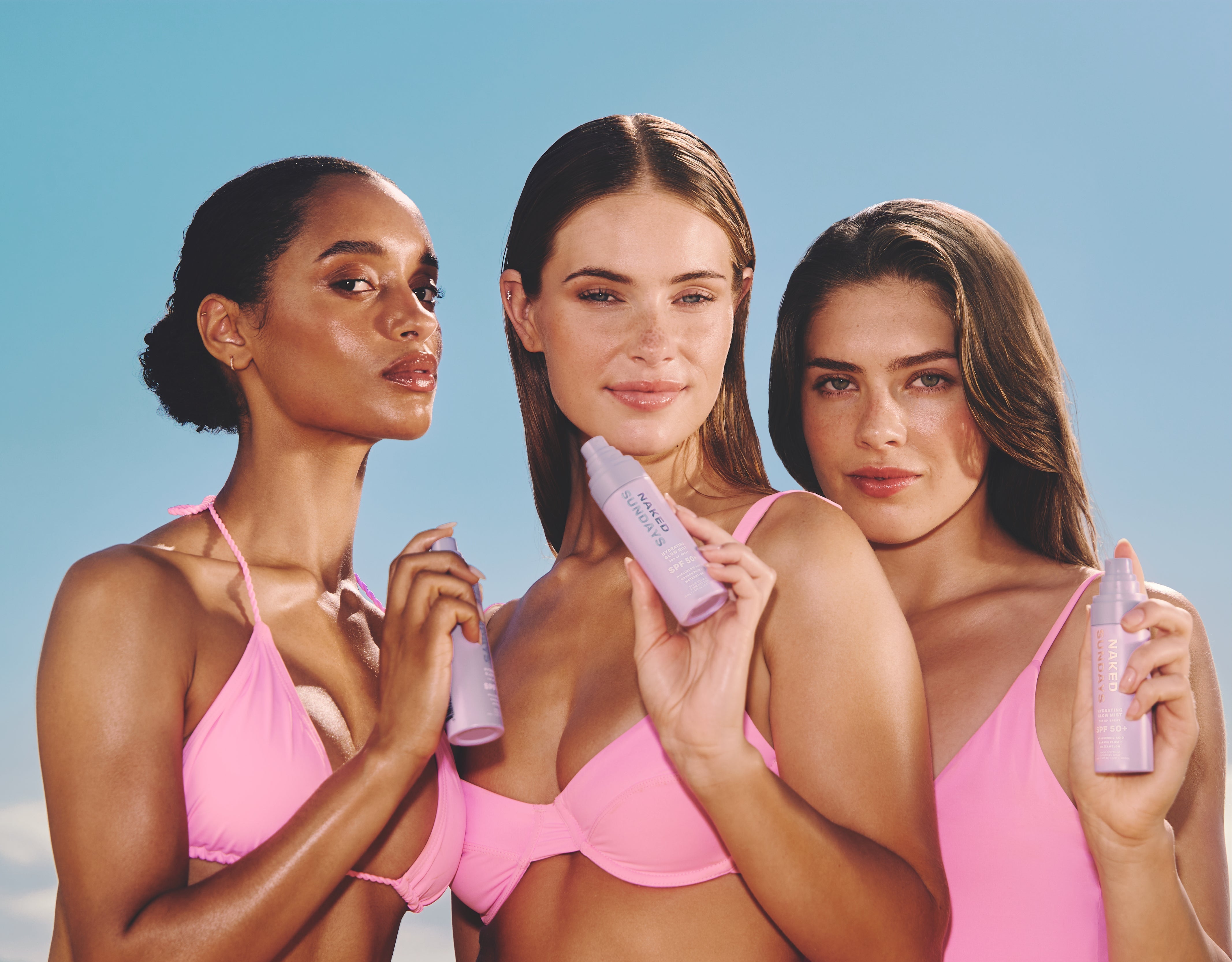Three women in pink bikinis holding spray bottles against a clear blue sky