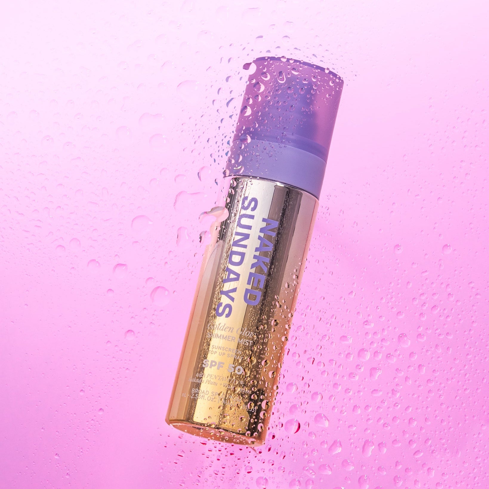 A bottle of Naked Sundays Golden Glow Shimmer Mist SPF50 on a pink background with water droplets surrounding the bottle