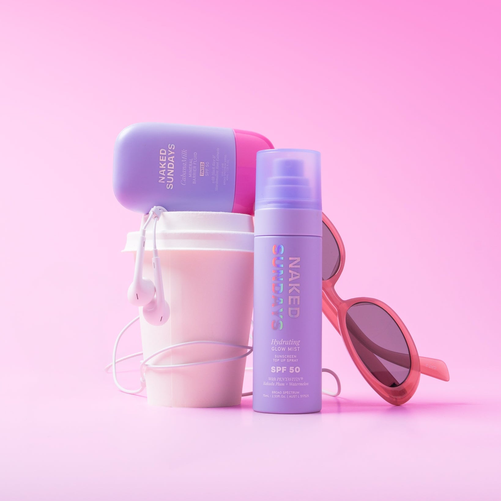 A styled product arrangement on a pink gradient background featuring a Naked Sundays Hydrating Glow Mist SPF 50 bottle, Naked Sundays CabanaMilk SPF50 priming fluid a white coffee cup with earphones,  and pink sunglasses