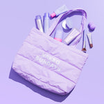 A lavender puffer tote bag with 'NAKED SUNDAYS' printed in white on the front, filled with various cosmetic items, photographed on a solid lavender background