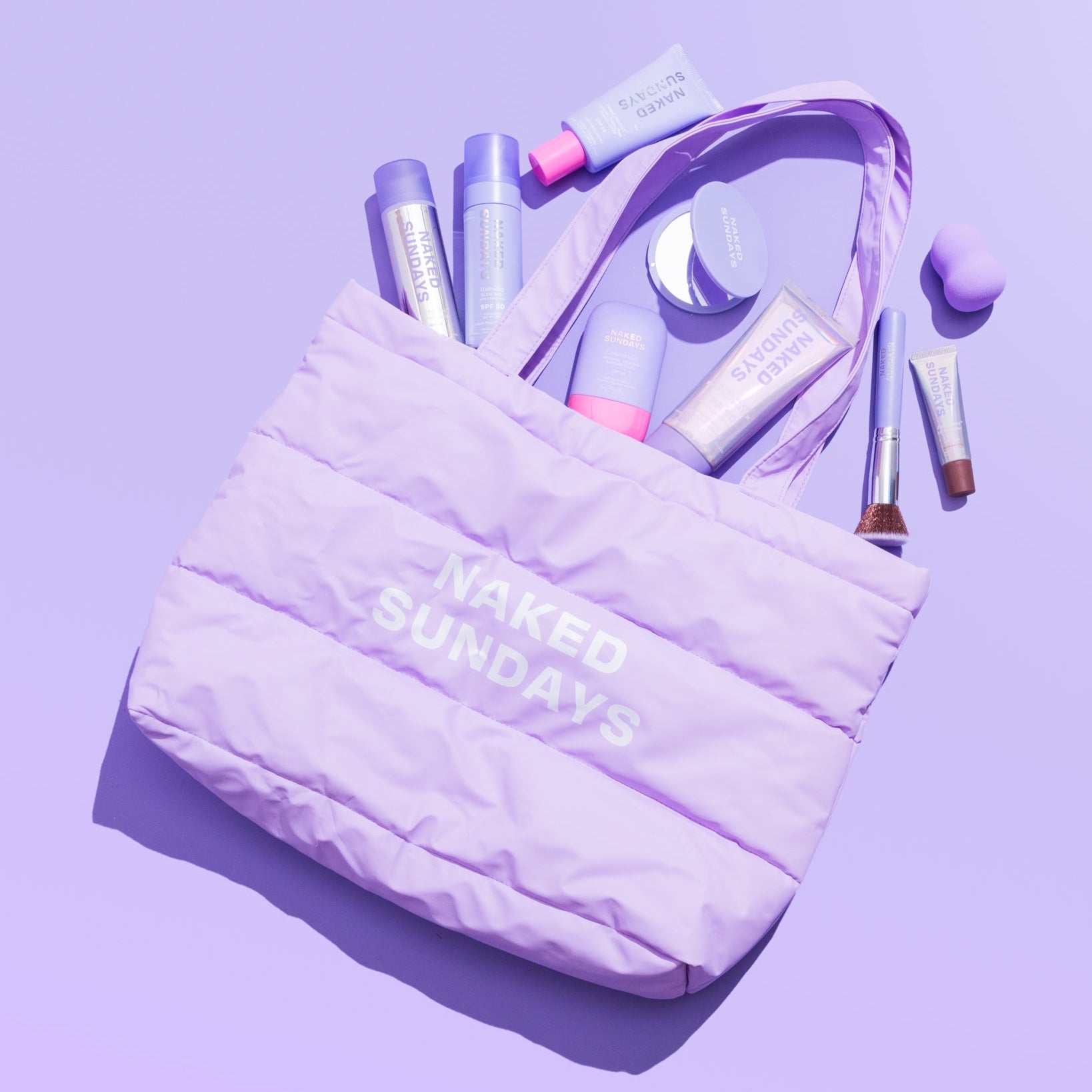 A lavender puffer tote bag with 'NAKED SUNDAYS' printed in white on the front, filled with various cosmetic items, photographed on a solid lavender background